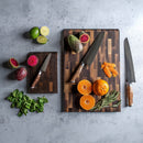 Two Steelport Knife Co. Signature Knife & Board Sets are displayed on a gray surface, each featuring American-made kitchen knives from Steelport Knife Co., along with oranges, prickly pears, lime halves, mint, and rosemary. Some fruits are expertly sliced using the handcrafted knives, while fresh herbs are artfully arranged alongside the boards.
