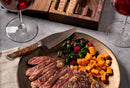 A plate of sliced steak, sautéed greens, and diced sweet potatoes is served with the Steelport Knife Co. Steak Knife Set, featuring handcrafted carbon steel steak knives from Steelport Knife Co. One of the knives rests on the plate, while two glasses of red wine and a wooden cigar box sit nearby on the gray table.