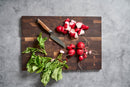 A Steelport Knife Co. SteelCore™ 2-in-1 Walnut Cutting Board, 18"x12", with a bunch of whole radishes, some sliced radishes, leafy greens, and a knife sits on a gray countertop.