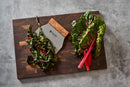 The Steelport Knife Co. SteelCore™ 2-in-1 Walnut Cutting Board, 18"x12", is displayed on a gray surface alongside a vegetable chopper. Chopped rainbow chard is neatly arranged on the left side of the board, while unchopped rainbow chard with vivid red stems sits on the right—showcasing both the practical design and stylish appeal of this steel-reinforced cutting board by Steelport Knife Co.