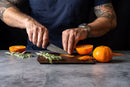 A person with tattoos slices orange peel on the Steelport Knife Co. SteelCore™ 2-in-1 Walnut Cutting Board (9"x6"), surrounded by whole and halved oranges and fresh herbs, on a gray countertop.