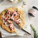 A rustic galette with prosciutto and rosemary sits beside a jar of Terrapin Ridge Farms Strawberry & Fig Jam, ideal for a cheese board. A glass of white wine and scattered rosemary sprigs complete the inviting scene.