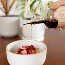 A hand pours Under The Bark Syrup Co Vanilla Maple Syrup onto yogurt topped with strawberries, set on a wooden table with potted plants in the background.