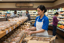 A grocery store worker in a blue shirt and white apron arranges individually packaged desserts and bulk pecan cakes, including the Texas Pecan Cakes Texas Pecan Cake 30-Pack Bulk Case for events and gatherings, on a display shelf. In the background, shoppers browse near the produce section and other grocery items.