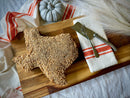 A Texas Pecan Cakes Large Texas Pecan Cake, crafted in the iconic shape of the Lone Star State and inspired by the classic Texas pecan cake recipe, rests on a wooden board sprinkled with chopped nuts. The scene is accented with a napkin, serving utensils, dried grass, a gray pumpkin, and a white cloth with red stripes—making this signature treat from Texas Pecan Cakes perfect for serving 8–10 and available for nationwide shipping.