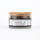 A glass jar with a wooden lid from Texas Salt Co. holds Texas Salt Co. Bourbon Barrel Smoked Vanilla Finishing Salt. The label shows the product name, weight, and ingredients: smoked salt and ground vanilla bean.