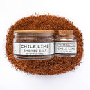 Two jars of Texas Salt Co. Chile Lime Smoked Salt, one large and one small with a cork lid, sit in front of a pile of pecan smoked salt. Both jars display white labels and are from Texas Salt Co.