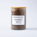 A glass jar with a wooden lid labeled Texas Salt Co. Cowboy Salt® (Net Wt 9oz/255g) contains coarse, brownish finishing salt from Texas Salt Co, shown against a white background.