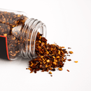 A glass jar of Texas Salt Co. Pecan Smoked Crushed Red Pepper lies on its side, with the spicy blend spilling out onto a white surface.