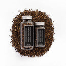 A large and a small glass jar of Texas Salt Co. Thick Hickory Smoked Peppercorn, both labeled in black and white, are shown atop a pile of loose smoked peppercorns.