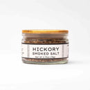 A small glass jar with a golden lid contains Texas Salt Co. Hickory Smoked Salt. The front label reads "Hickory Smoked Salt, Net Wt 2.75oz (78g)" in bold black letters—perfect for adding delicious BBQ smoke flavor to your favorite dishes.