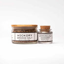 Two jars of Texas Salt Co. Hickory Smoked Salt are pictured: the large jar has a gold lid, the small one a cork lid. Both display the Texas Salt Co. label in black on white, highlighting their rich BBQ smoke flavor.