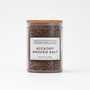 A clear glass jar with a wooden lid holds Texas Salt Co. Hickory Smoked Salt, adding rich BBQ flavor. The label reads: Texas Salt Co. Hickory Smoked Salt Net Wt 7oz (198g). Set against a plain white background.