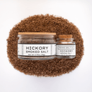 Two glass jars of Texas Salt Co. Hickory Smoked Salt, one large and one small, sit on a white background with coarse smoked sea salt spilled beneath them. Both jars have white labels and wooden lids—perfect for adding BBQ flavor.