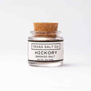 A small glass jar with a cork lid labeled Texas Salt Co. Hickory Smoked Salt, Net Wt. .5oz (14g), from Texas Salt Co., adds bold BBQ smoke flavor to your dishes and is shown on a plain white background.