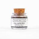 A small glass jar with a cork lid labeled Texas Salt Co. Jalapeño Smoked Salt, Net Wt. .75oz (21g), makes a bold finishing salt and is shown on a white background.