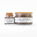 Two Texas Salt Co. Jalapeño Smoked Salt jars—one small with a cork lid, one large with a wooden lid—are displayed side by side on a white background. Both feature white labels with black text, making them perfect as finishing salts.