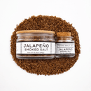 Two glass jars of Texas Salt Co. Jalapeño Smoked Salt—one large with a wooden lid, one small with a cork top—sit artfully arranged, both labeled and perfect for adding bold flavor to your dishes.