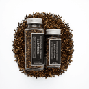 Two glass jars labeled Texas Salt Co. Mesquite Smoked Peppercorn sit on a pile of loose smoked black pepper against a white background. The jars, in different sizes with silver lids, highlight this bold Texas Salt Co. smoked seasoning.