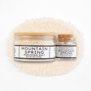 A large and small jar of Texas Salt Co. Mountain Spring Peruvian Salt, both with wooden lids and white labels, sit atop coarse Andean salt, highlighting the product name and weight.