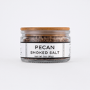 A clear glass jar with a wooden lid holds Texas Salt Co. Pecan Smoked Salt. The white label features “Pecan Smoked Salt, Net Wt 3 oz (85g)” in bold black text and the Texas Salt Co brand name.