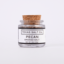 A small glass jar with a cork lid labeled “Texas Salt Co. Pecan Smoked Salt” shows Texas Salt Co.’s pecan smoked salt, net wt 0.5oz (14g), on a plain white background.
