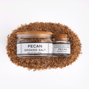 A large and a small glass jar of Texas Salt Co. Pecan Smoked Salt, both with white labels and cork lids, sit on a mound of brown smoked salt. The jars feature Texas Salt Co branding, all set against a bright white background.