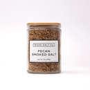 A glass jar with a wooden lid, labeled Texas Salt Co. Pecan Smoked Salt, holds coarse brown pecan smoked salt (net wt 7 oz/198g) by Texas Salt Co. The jar is set against a plain white background.