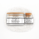 Two glass jars of Texas Salt Co. Republic of Senegal African Salt—one large with a wooden lid and one small with a cork stopper—are displayed on coarse white salt crystals.