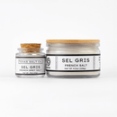 Two jars of Texas Salt Co. Sel Gris French Salt. The smaller jar has a cork lid, and the larger one a wooden lid. Both have minimalist white labels with weight info, featuring the Texas Salt Co brand name.