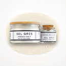 Two glass jars of Texas Salt Co. Sel Gris French Salt sit on a mound—one large with a wooden lid, the other small with a cork. Both white labels display the product name, showcasing this mineral-rich sea salt’s authentic French origin.