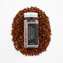 A clear jar labeled "Texas Salt Co. Szechuan Peppercorn" from Texas Salt Co. lies on its side atop a circular pile of loose red peppercorns, a vibrant ingredient in Sichuan cuisine, on a white background.