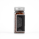 A clear glass jar with a silver lid, labeled Texas Salt Co. Szechuan Peppercorn, Net Wt: .75 oz (21g), contains Szechuan peppercorns essential for Sichuan cuisine and sits on a white background.