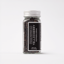 A clear glass jar with a silver lid, filled with premium whole peppercorns. The label reads Texas Salt Co. Tellicherry Peppercorn and shows a net weight of 2 oz (57 g). The background is white.