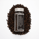 A clear bottle labeled Texas Salt Co. Tellicherry Peppercorn sits on loose whole peppercorns, showing premium peppercorns inside. The label also displays Texas Salt Co. and Net Wt. 2 oz (57g).