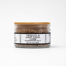 A clear glass jar with a wooden lid, labeled Texas Salt Co. Tequila Barrel Smoked Lime Finishing Salt, 3oz (85g), filled with coarse brown tequila barrel smoked lime salt, on a white background. Adds savory citrus smoke to any dish.