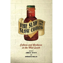 A paperback copy of "The Slaw and the Slow Cooked: Culture and Barbecue in the Mid-South" from Books by splitShops is pictured with a ribbon label, set against a brick wall backdrop. Edited by James R. Veteto and Edward M. Maclin, this book delves into the traditions and culture of mid-South barbecue within America's rich culinary history.