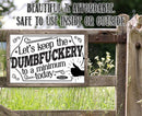 A wooden fence displays the Let’s Keep The Dumbfuckery - Metal Sign by Lone Star Art—an irreverent, stylish, and affordable gift for indoor or outdoor spaces. Green grass and plants complete the scene.