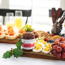 Terrapin Ridge Farms Hot Pepper Bacon Jam is displayed on a wooden board with deviled eggs, grapes, berries, and appetizers—ideal for charcuterie boards—alongside drinks and food on a holly-themed table.