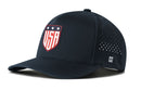 Introducing the Haka Hat USA Shield Performance 5-Panel Hat | America Flexfit Snapback by Haka Hat—a dark blue performance hat featuring a red, white, and blue shield logo on the front, small white ventilation holes on the side for breathability, and a subtle white H logo near the brim. This water repellent snapback is equipped with a comfortable Flexfit sweatband to keep you cool and dry throughout the day.