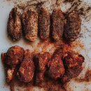 Eight pieces of seasoned chicken, generously coated in a rich blend of dark spices from the Dead Rooster Co. Variety Pack - 3 Signature BBQ Rubs Bundle & Save, are arranged in two rows on a white surface with extra rub from Dead Rooster Co. sprinkled around.
