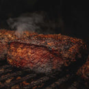 A close-up of a thick, juicy steak with a charred crust, sizzling on the grill as smoke rises in the background—made irresistible by the bold flavors from the Dead Rooster Co. Variety Pack - 3 Signature BBQ Rubs Bundle & Save.