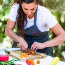 During the Messermeister HQ Cooking Class by Messermeister, a woman in a white shirt and dark apron slices colorful bell peppers on a wooden cutting board outdoors, surrounded by fresh vegetables.