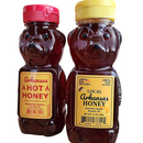 Two bear-shaped bottles of honey sit on a table. The left, with a red cap, is labeled Hot Honey; the right has a yellow cap and is Guenther Apiary Arkansas Wildflower Honey Bear by Guenther Apiary.