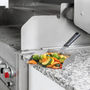 A Fox Run Brands XL Stainless Steel Griddle Shovel – 5 Cup Food Scoop & Spatula filled with sautéed vegetables rests on a granite countertop next to a grill.