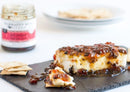 A block of cheese topped with Terrapin Ridge Farms Hot Pepper Bacon Jam is served on a slate with crackers—ideal for charcuterie boards. In the background, a jar of Terrapin Ridge Farms bacon jam and a plate of crackers are blurred.