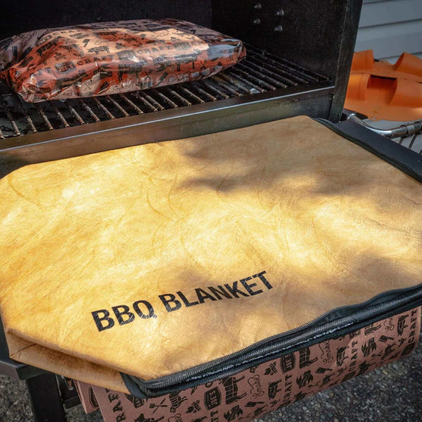 A tan Drip EZ BBQ Meat Resting Blanket, labeled Drip Ez, is draped over a grill lid, with another insulated meat resting blanket inside to lock in juices. Orange BBQ accessories can be seen in the background.
